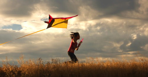 Young girl running in wheat field with kite under cloudy skies
