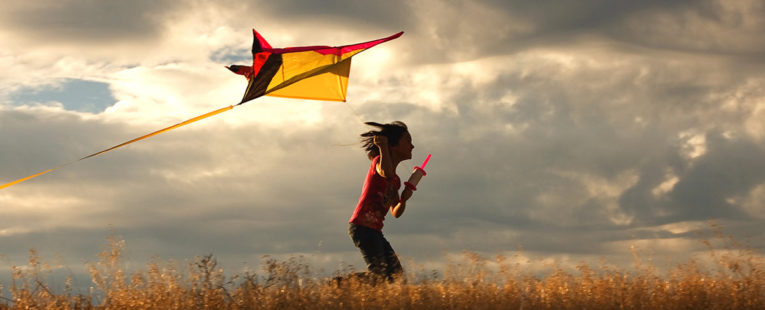 Young girl running in wheat field with kite under cloudy skies