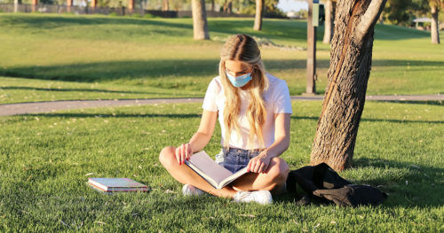 Young masked woman college student reading textbook outdoors.