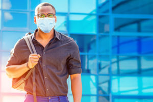 Young masked African-American male college student outside campus building.