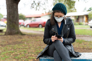 Young masked woman college student reading her phone in a park.