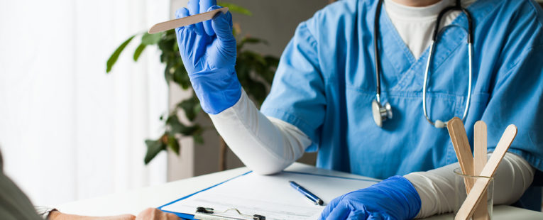 Doctor at desk in face mask and gloves with tongue depressor; patient hands visible in foreground