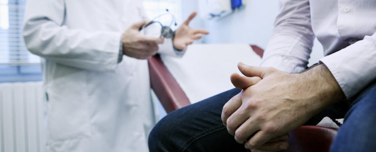 Man sits in foreground on table in doctor's office, only visible from mid-chest to knees, with doctor in background, hands out as though emphasizing a discussion point
