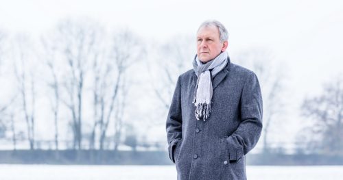 senior man stands solemnly outside in a coat and scarf during winter