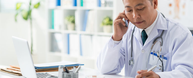 Male Asian doctor at desk speaking on phone with notepad and laptop in front of him