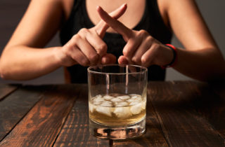 woman behind an glass of alcohol with ice, fingers crossed as if to say "no"