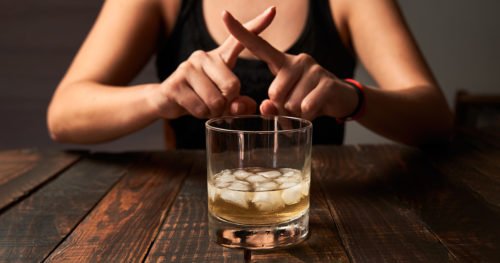 woman behind an glass of alcohol with ice, fingers crossed as if to say "no"