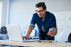 man leans over the table typing on his laptop while simultaneously holding his cellphone