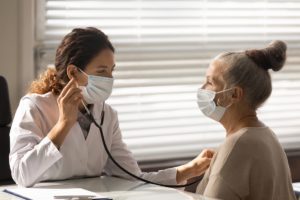 masked female doctor uses a stethescope on a senior woman in an exam room