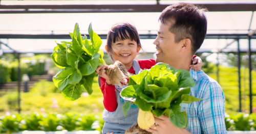 father holds his daughter and both are holding freshly picked vegetables