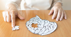 Elderly woman hands putting missing white jigsaw puzzle piece down into the place as a human brain shape. Creative idea for memory loss, dementia, Alzheimer's disease and mental health concept.