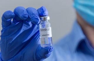 Medical professional wearing blue collared shirt and blue surgical mask in background holds up vaccine vial in foreground between two fingers while wearing blue latex gloves