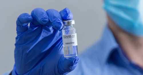 Medical professional wearing blue collared shirt and blue surgical mask in background holds up vaccine vial in foreground between two fingers while wearing blue latex gloves