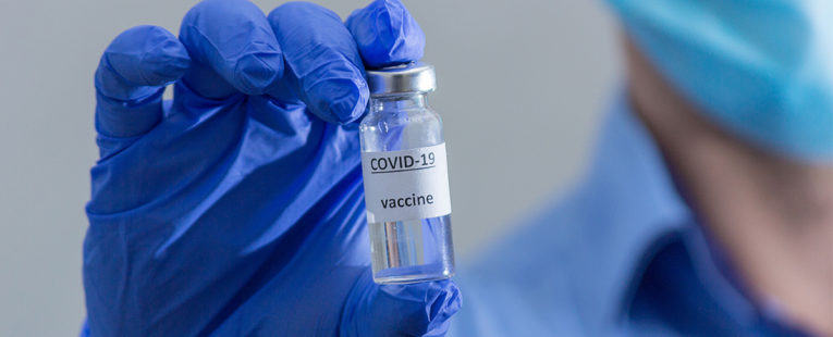 Medical professional wearing blue collared shirt and blue surgical mask in background holds up vaccine vial in foreground between two fingers while wearing blue latex gloves