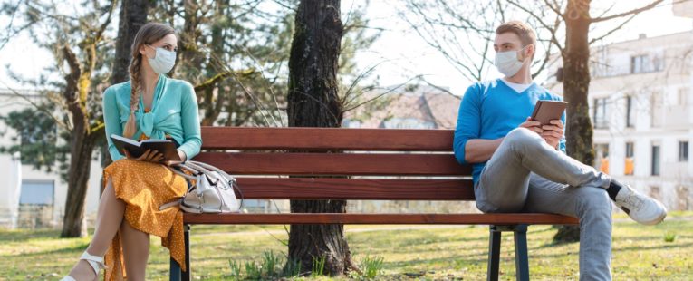 Two people with masks sit far apart from each other on a bench to help prevent the spread of COVID.