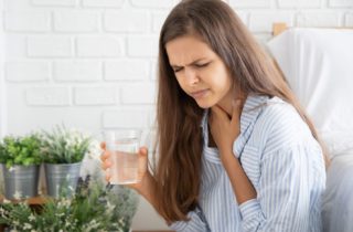 Long-haired brunette woman holds glass of water in right hand and throat in left hand with concerned look on face as though she is experiencing hiccups