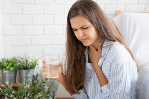 Long-haired brunette woman holds glass of water in right hand and throat in left hand with concerned look on face as though she is experiencing hiccups