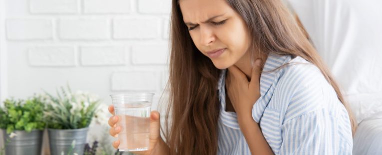 Long-haired brunette woman holds glass of water in right hand and throat in left hand with concerned look on face as though she is experiencing hiccups