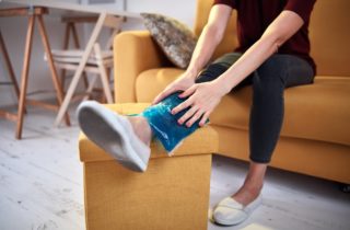 Individual in black workout clothes sits on yellow couch with leg extended onto a yellow ottoman, where she holds a small ice bag to her ankle