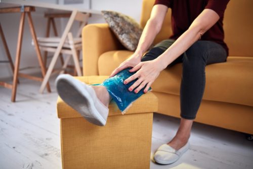 Individual in black workout clothes sits on yellow couch with leg extended onto a yellow ottoman, where she holds a small ice bag to her ankle