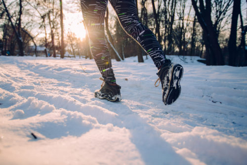 Sun shines in the background of a wooded area as a runner runs through the snow in the foreground