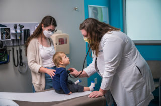 A woman and child visit an OnCall Urgent Care provider. The provider is checking the child's heart with a stethoscope.