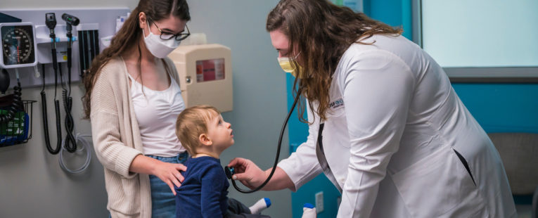 A woman and child visit an OnCall Urgent Care provider. The provider is checking the child's heart with a stethoscope.