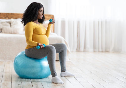A pregnant woman sits on an exercise ball and holds weights to safely work out during pregnancy.