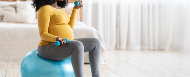 A pregnant woman sits on an exercise ball and holds weights to safely work out during pregnancy.