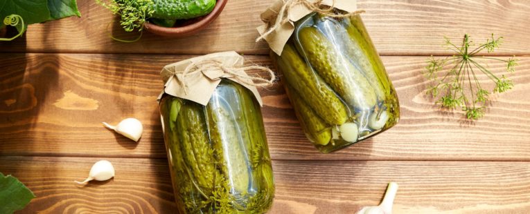 Pickled cucumbers in jars, fresh cucumbers on bowl, dill, garlic on a wooden background. Home canning. Top view.