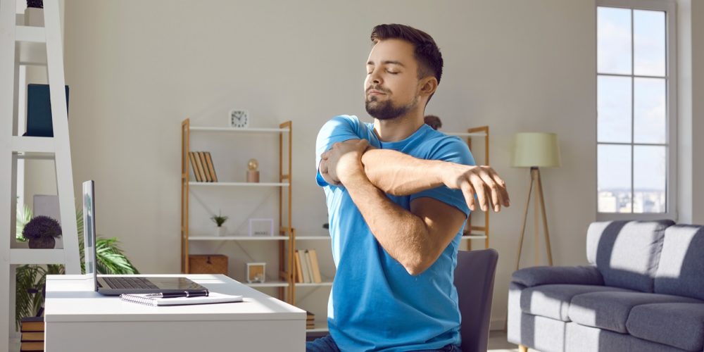 young bearded man stretches his arm across his chest sitting at his desk