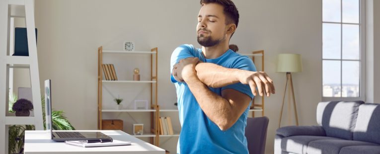 young bearded man stretches his arm across his chest sitting at his desk